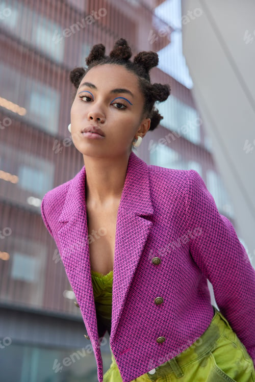 Preview: Attentive fashionable woman with bun hairstyle looks seriously at camera dressed in stylish pink jac