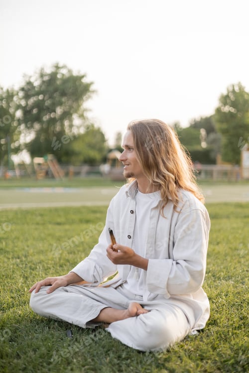 Preview: carefree man in white cotton clothes meditating in easy pose with fragrant palo santo stick