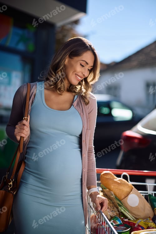 Preview: Satisfied pregnant woman with shopping cart after shopping at supermarket.