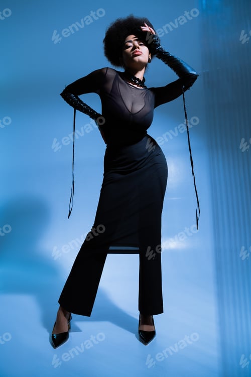 Preview: full length of curly african american model in black outfit posing with hand on hip on blue