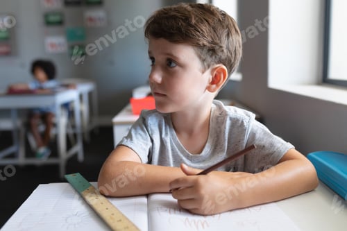 Visualização: Estudante primário caucasiano com livro e lápis olhando para longe enquanto está sentado à mesa na sala de aula