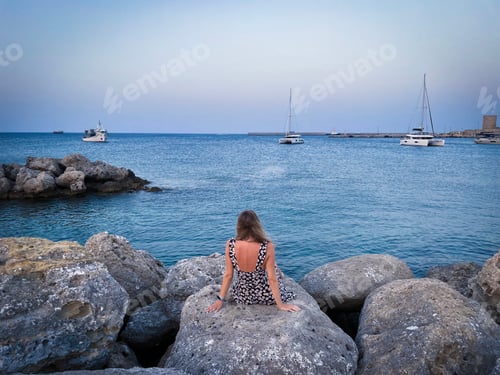 Preview: Rear view of woman sitting down on the cliffs looking at the ships sailing the blue sea in Greece