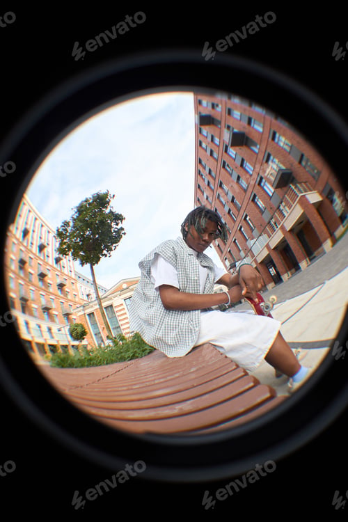 Preview: Young Black Man Sitting Outdoors Adjusting Skateboard in Urban Setting