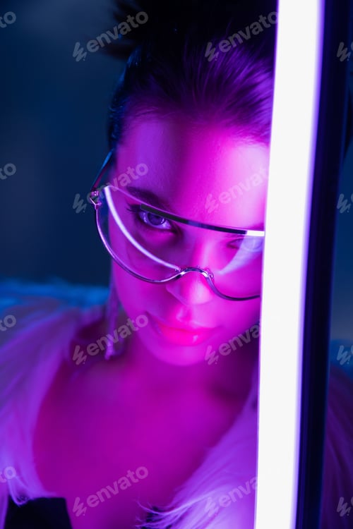 Preview: Trendy young woman in sunglasses looking at camera near lamp on navy blue background