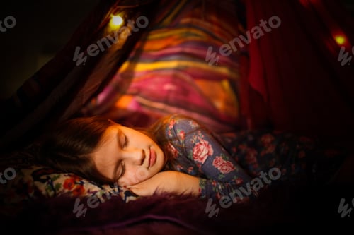 Preview: Caucasian kid girl sleeping in a toy tent with garlands, children's games and houses, dark cozy phot
