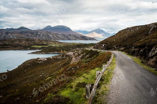 Preview: Landscape with rural road cutting through mountains and lochs under a cloudy sky.
