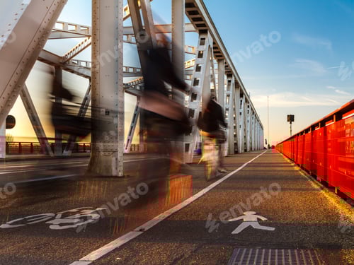 Preview: Ghosts cycling on cycle path crossing bridge long exposure blurred people on bike. Low sunlight glow