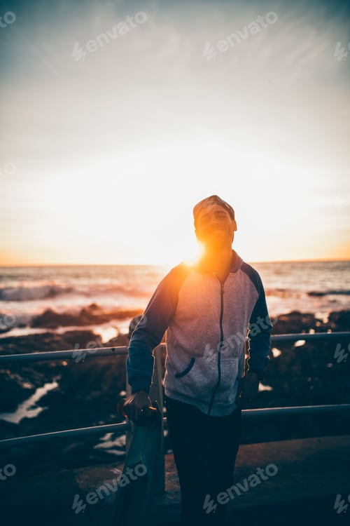 Preview: Skater boy with longboard in sunset on promenade