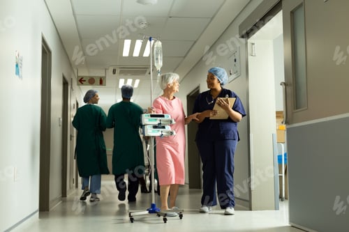 Preview: Happy diverse female doctor and senior female patient with iv pole talking in busy hospital corridor