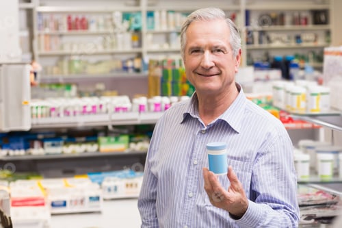 Preview: Senior male pharmacist holding medicine container at pharmacy counter with shelves and dispenser