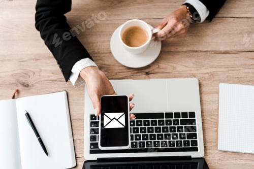 Preview: top view of businesswoman holding smartphone with envelope on screen and cup with coffee near laptop