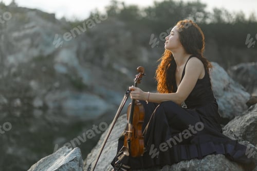 Preview: Girl in black dress and curly hair violinist sitting on Rocky Mountain with real vintage violin.