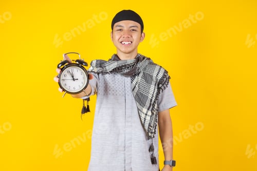 Preview: Smiling Young Man in Muslim Clothes Holding Vintage Alarm Clock on Yellow Background