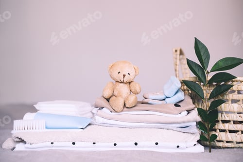 Preview: Pile of clean baby clothes, skin care cosmetic, teddy bear toy and laundry basket for newborn.