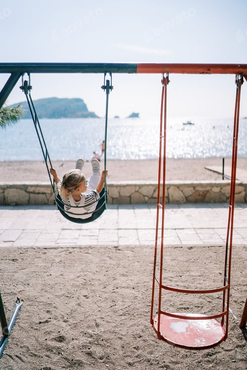 Preview: Little girl swings on a colorful swing at a playground by the sea, looking to the side. Back view