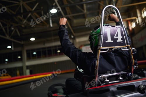 Preview: back view of motorsports driver in helmet raising hand and winning race on circuit, go-kart