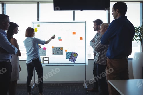 Preview: Businesswoman sticking adhesive notes on whiteboard while colleagues standing at office