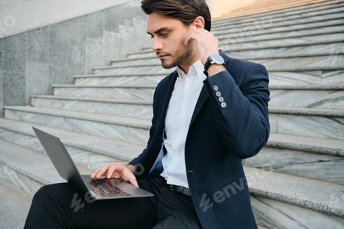 Preview: Young handsome man in classic suit sitting on stairs outdoor thoughtfully working on laptop