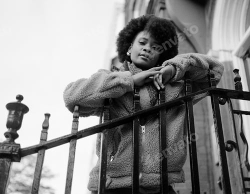 Preview: Analog portrait of young black woman leaning on bar banister outside building.