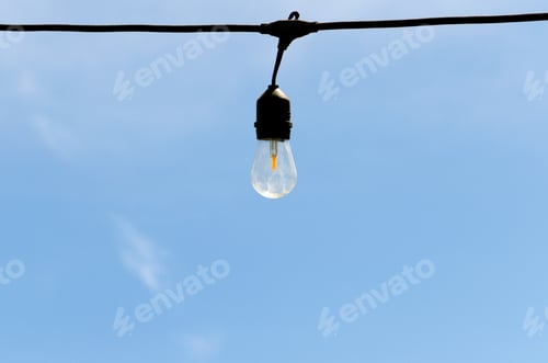 Preview: One single hanging Edison lightbulb on a wire against a bright blue sky with some clouds, minimal