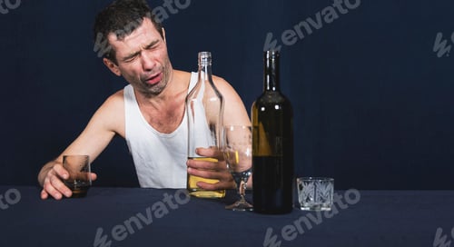 Preview: Drunk lonely man in home jersey sits at table, holds bottle of alcohol in his hand and looks at it