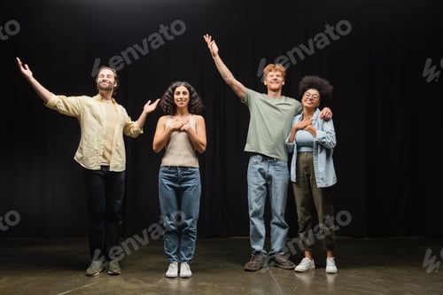Preview: full length of happy multiethnic actors standing with raised hands on scene of theater