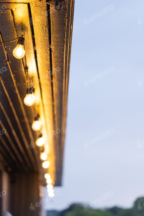 Preview: Close-up of warm glowing string lights hanging from a wooden roof at dusk