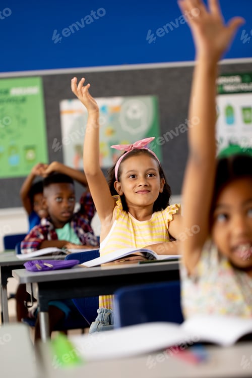 Preview: Diverse elementary schoolchildren sitting at desks and raising hands in elementary school class