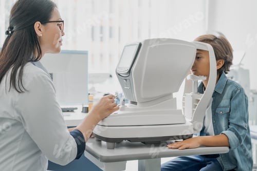 Preview: Side view shot of little boy having test for his eyes with special optical apparatus female doctor