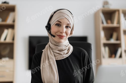 Preview: Smiling positive confident woman in hijab and headset sitting at office desk.