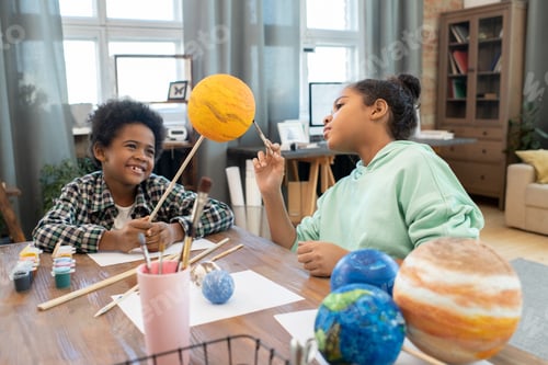 Preview: Two adorable mixed-race schoolkids in casualwear painting planets by table