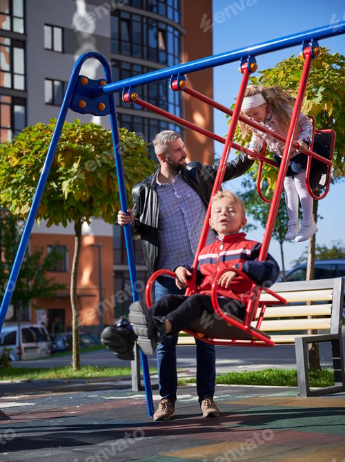 Preview: Dad swinging cute children on swings at playground in autumn.