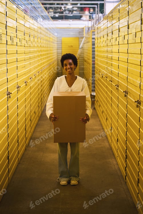 Preview: Happy young woman standing in warehouse with cardboard box