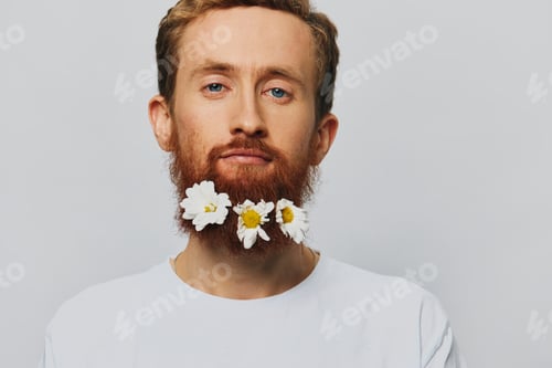Preview: Portrait of a funny man in a white T-shirt with flowers daisies in his beard on a white isolated