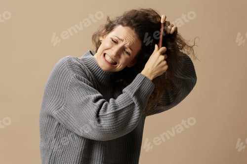 Preview: Suffering curly beautiful female in gray casual sweater with hairbrush comb pulls tangled hair