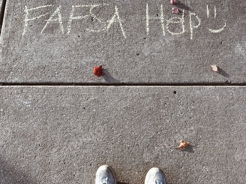 Preview: College student looking down at words on sidewalk written in chalk with message for FAFSA help