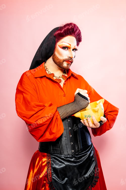 Preview: Drag Queen Dressed in Vibrant Attire Posing With Mortar Against Pink Background