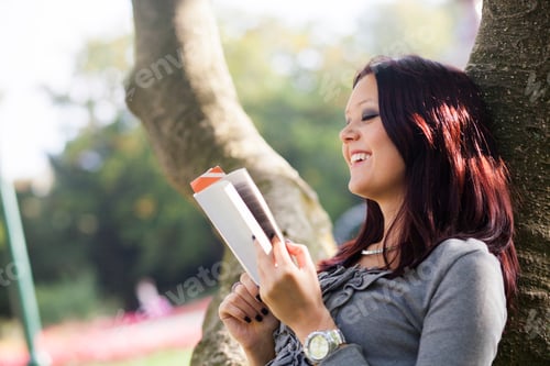 Preview: Happy female student reading book while resting on tree trunk at university campus