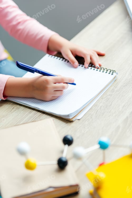 Preview: cropped shot of schoolchild taking notes while studying with books and molecular model