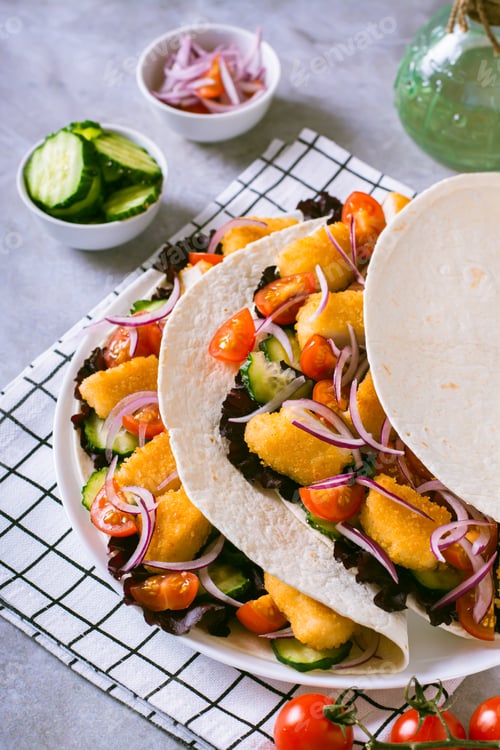 Preview: Tacos with chicken nuggets, cucumber, tomato, onion and lettuce on a plate vertical view