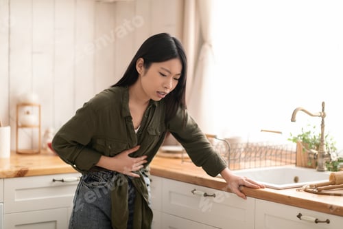 Preview: Young asian woman feeling sick in kitchen, touching her belly
