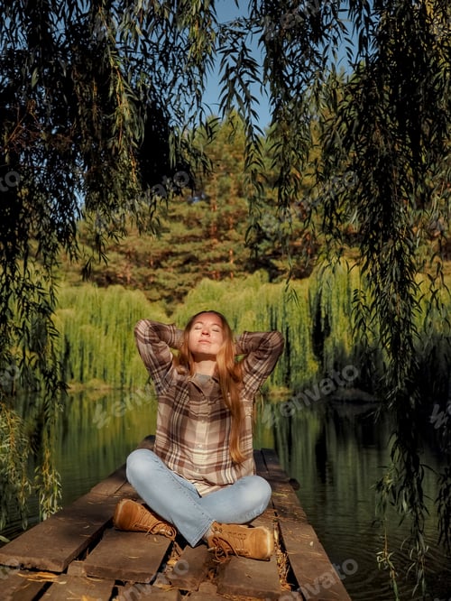 Preview: A girl in a checkered shirt sits on a bridge near the lake and meditates