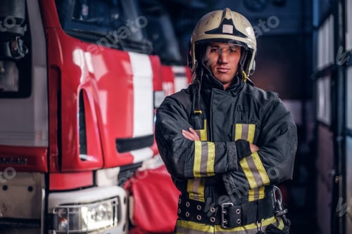 Preview: Fireman wearing protective uniform standing next to a fire engine in a garage of a fire department