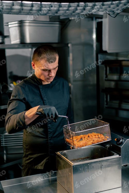Preview: the chef in the kitchen in a black jacket takes out baht from the deep fryer