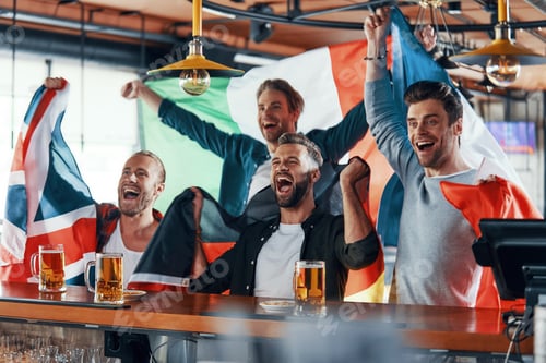 Preview: Cheering young men covered in international flags enjoying beer while watching sport game in the pub