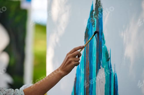 Preview: Artist hand holds paint brush and draws abstract surreal image on white canvas at outdoor festival