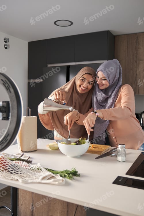 Preview: Two muslim food vloggers smiling while pouring olive oil in a salad at kitchen.