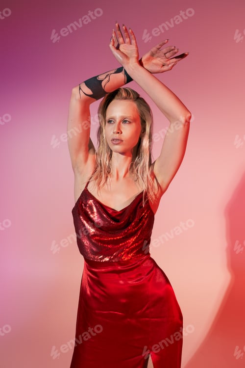 Preview: appealing young woman with tattoo in red chic dress posing with arms raised on pink backdrop