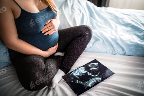 Preview: Pregnant woman sitting on bed and looking at ultrasound photo of her baby