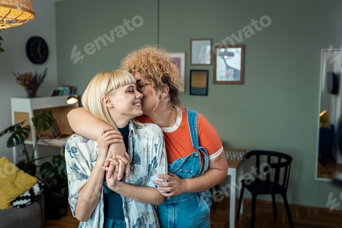 Preview: Portrait of a smiling lesbian couple standing in a room, kissing each other in cheek.